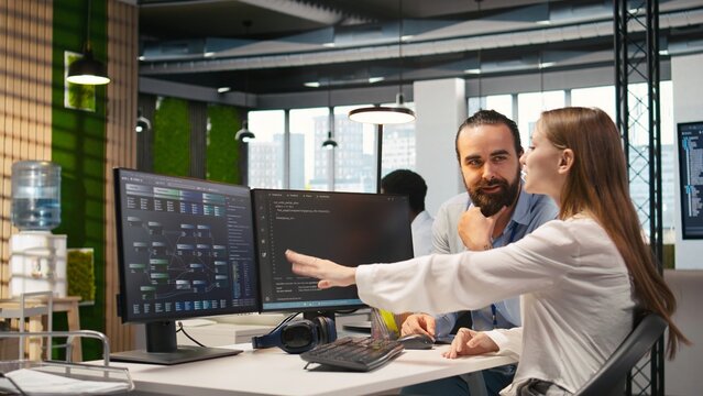 Friendly team leader helping happy young engineer code AI algorithms to debug software issues and improving system stability. Helpful manager assisting smiling intern in tech startup office, camera A