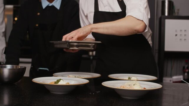 Cropped shot of male chef adding final touches to prepared dishes sprinkling food with grated cheese during group culinary tutorial in restaurant kitchen