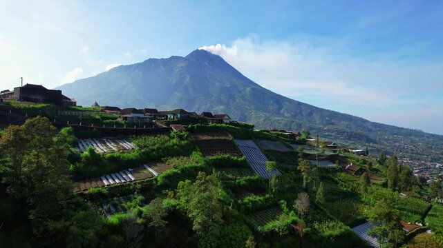 Stunning aerial drone footage of Merapi volcano mountain still smoking with ashes at sunrise, with a beautiful village and lush valley in the foreground