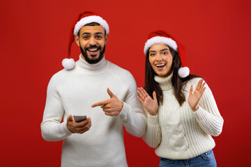 Fototapeta premium A cheerful Arab couple wearing Santa hats is happily surprised while receiving a Christmas offer on a smartphone. They are posing together in front of a red background.