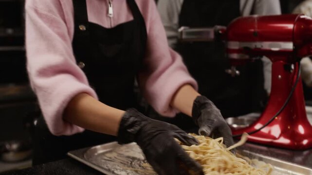 Tilt up shot of young woman dusting fresh artisanal pasta with flour tossing noodles on tray while groupmate observing during cooking class in restaurant kitchen