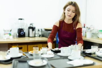 Diligent young waitress taking away dirty dish on trays in cozy cafeteria