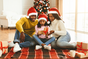 A family sits on the floor, wearing red hats, as they joyfully unwrap gifts. The room has a Christmas tree and presents surrounding them. The child appears excited to see what is inside a box.