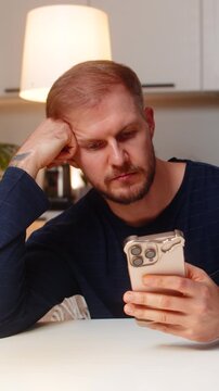 Young man at night home scrolls smartphone reads news feels boredom stress fatigue loses focus. Caucasian guy on sofa at table stares at phone quiet mood doomscrolling tired seeks rest tea mug near.