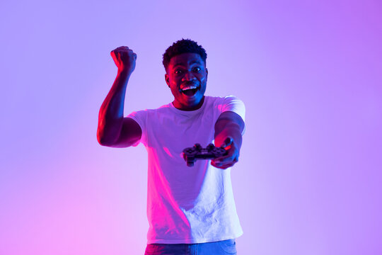An excited young black man cheers in celebration after winning a video game competition. He holds a joystick and gestures with joy amidst bright neon lights. - Powered by Adobe