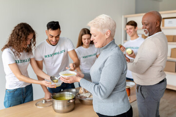 A happy senior woman smiles as she receives warm soup from young volunteers at a charity office. The group is dedicated to providing free meals for elderly people in need.