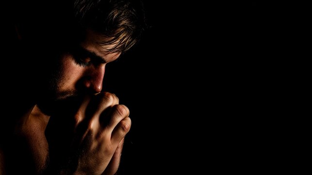 Young man praying with hands clasped in dark room with low key lighting  