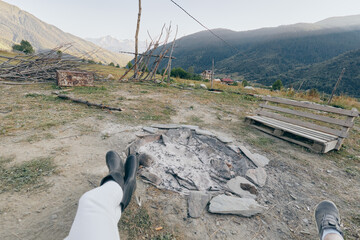 Fototapeta premium Campfire mountains boots bench firepit hiking outdoors relaxation. Two people resting with boots stretched toward a stone firepit and wooden bench, panoramic mountain valley view at campsite.