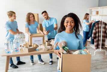 A young woman smiles while holding a box of toys for donation. Behind her, diverse volunteers work together, packing clothes and food in boxes at a charity center.
