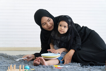 Happy Muslim mother and cute little daughter playing with cooking toys on floor at home. Islamic family lifestyle concept with parent and kid enjoying activity together on carpet.