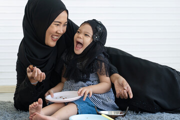 Cheerful Muslim mother and cute daughter eating dates fruit together on floor at home. Happy Islamic family lifestyle concept during Ramadan Iftar or snack time with laughter.