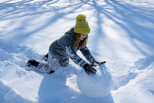 Smiling child building a snowman with snow on a sunny winter day. Happy girl playing in snow outdoors. Cute kid making a snowman. Child paly wit snow. Kid building a big snow ball in winter field.