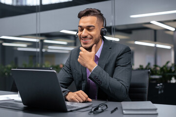 A man in formal attire sits at a desk in an office. He wears headphones and looks at a laptop. He has a pleasant expression on his face while engaging in a discussion.