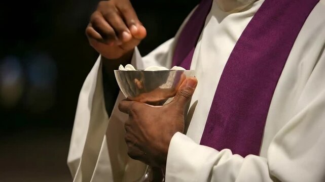 Hands of a priest holding a silver bowl containing sacramental bread or communion wafers