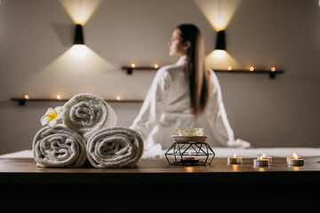Relaxing spa setting featuring neatly rolled towels, aromatic candles, and a serene woman in a white robe, creating a tranquil atmosphere for massage therapy and wellness