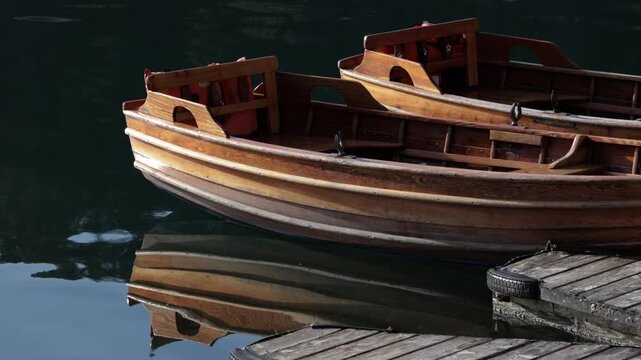 Close-up of a small, narrow brown wooden boat docked perpendicular to a wooden pier, with its reflection visible in gentle ripples on the calm water.