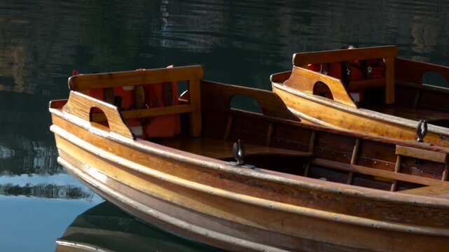 Close-up of a small, narrow brown wooden boat docked perpendicular to a wooden pier, with its reflection visible in gentle ripples on the calm water.