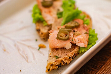 Close-up of smoked salmon open sandwich on seeded bread with lettuce and sliced jalapeno served on a white plate. Gourmet appetizer and café food concept