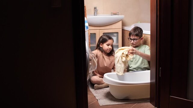 Young boy and girl sitting on the floor, loading clothes in a white basin in the bathroom, actively contributing to household chores and learning responsibility