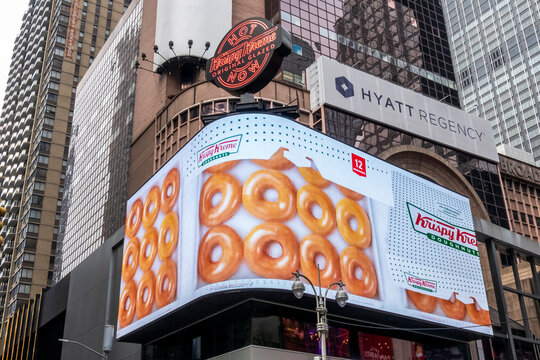 New York, NY - US - Dec 13, 2025 A busy Times Square street scene featuring the brightly lit Krispy Kreme flagship store and its massive corner-wrapping LED display showing glazed doughnuts.