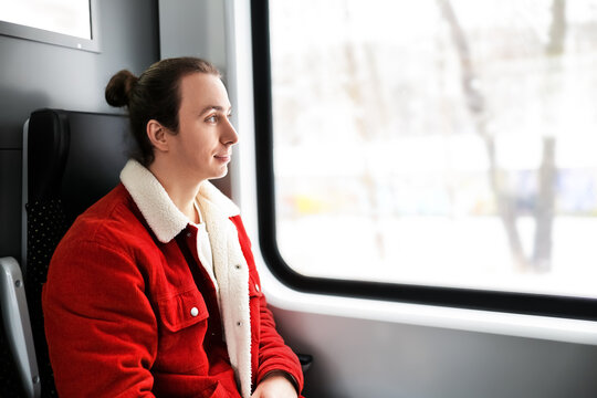  man with bun hairstyle in a red jacket sitting on a train and looking thoughtfully out the window at winter scenery.