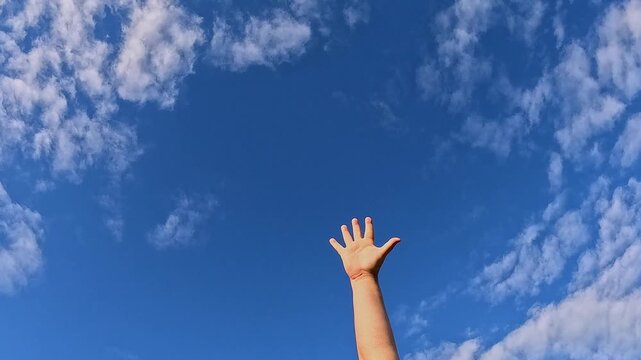 Single human arm and hand reach up from bottom frame, open palm, spread fingers and wave gently as a gesture under bright blue sky while small white clouds drift past, expressing freedom and hope.