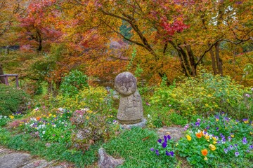 満開のカラフルな花とちょうど見ごろの紅葉に囲まれて微笑むかわいいお地蔵様