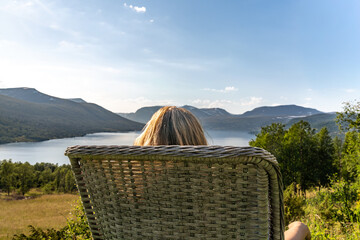 A woman sits on a chair in a garden, watching Gjevillvatnet lake surrounded by mountains in Oppdal Norway. She enjoys her free time in the summer sun and fresh air.
