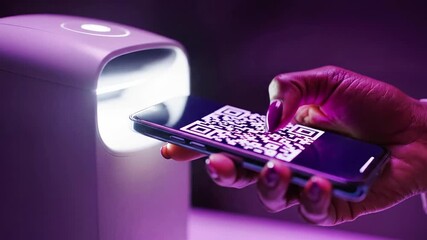 A hand holding a smartphone with a QR code on the screen next to a purple-lit device - Powered by Adobe