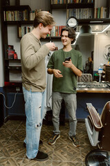 Two friends laugh and share a moment in a cozy barbershop. One holds a drink and the other checks...