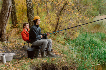 Two friends sit at the riverbank, surrounded by vibrant autumn colors, casting their fishing lines...