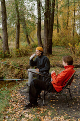 Two friends relax by a tranquil lake surrounded by colorful autumn leaves. One is sipping a drink while the other prepares to cast a fishing line. Laughter and nature fill the air.