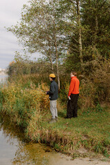 Two friends stand at the edge of a serene lake, surrounded by colorful autumn foliage. They admire the tranquil water and share a quiet conversation as leaves fall gently around them.