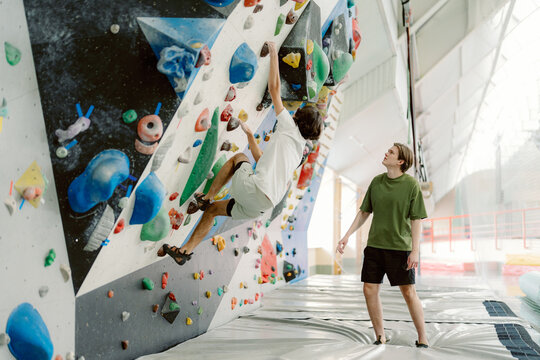 Two friends enjoy an exciting day at an indoor climbing gym. One is mid-climb on the colorful wall while the other provides encouragement from below, creating a fun atmosphere.
