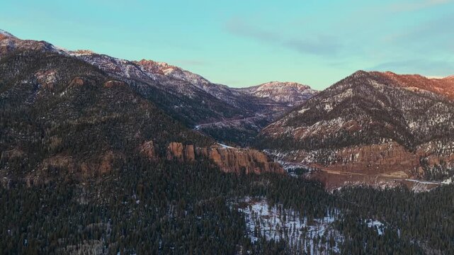 Wide drone view of Highway 160 carved into snowy cliffs during golden hour. A vehicle travels along the winding mountain road through a dense pine forest in the San Juan Mountains.