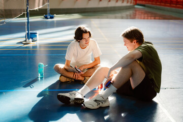 Two young men sit on the gym floor, sharing laughs and stories after an intense basketball session. The sunlight streams through the windows, creating a warm atmosphere.