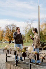 Two friends stand outside in a vibrant autumn park, sharing drinks and laughter. Golden leaves surround them as they engage in a warm conversation, creating a joyful atmosphere.