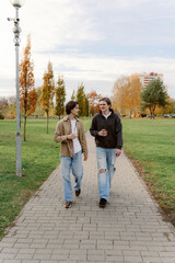Two friends stroll through a picturesque park, surrounded by colorful autumn trees. They share laughs and drinks, enjoying the crisp air and beautiful scenery together.