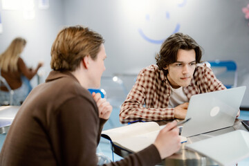 Two young men sit closely at a table in a vibrant cafe, focused on a laptop screen. One takes notes while the other types, showcasing teamwork on a study project.