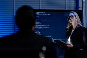 A female software architect presents the system architecture or walks through a complex codebase on a large screen using a tablet to guide her development team during a review.