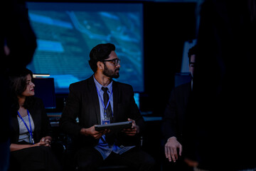 An Indian male data scientist and an Indian female engineer attend a tech conference. They are watching a presentation about AI and data analytics in a control room.