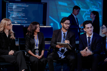 A large, diverse team of business professionals in a group meeting. An Indian man with a tablet leads the discussion with Asian and Caucasian colleagues in a high-tech office.
