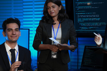 An Indian female developer leads a code review, holding a report. She explains a Python script for an AI or cybersecurity system to her team in a dark SOC Security Operations Center.
