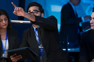 An Indian data scientist explains an AI algorithm on a tablet to a Caucasian software engineer. They are reviewing system data in a cybersecurity command center