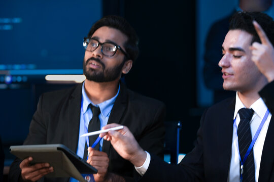 An Indian data scientist explains an AI algorithm on a tablet to a Caucasian software engineer. They are reviewing system data in a cybersecurity command center - Powered by Adobe