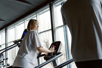 Fototapeta premium A woman happily uses an elliptical trainer in a spacious gym filled with natural light. Nearby, another person focuses on their workout. The atmosphere is energetic and motivating.