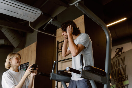 A man stands on a fitness machine, covering his ears while a woman assists him with a device. They are in a modern gym with wooden accents and motivational quotes on the walls. - Powered by Adobe