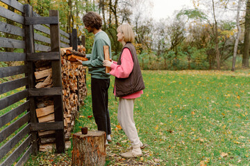 In a peaceful outdoor space filled with fallen leaves, a couple works together to stack firewood. The man organizes logs while the woman assists from behind, enjoying their time outdoors.