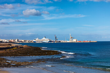 Port of Puerto del Rosario, capital of Fuerteventura in Canary Islands. Foreground features rocky coastline leading to harbor with large commercial cranes and a cruise ship docked at the pier
