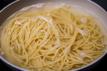 An overhead, close-up shot of spaghetti pasta actively cooking in a pot of boiling water. The noodles are fully submerged, and small bubbles and froth are visible on the water's surface.
